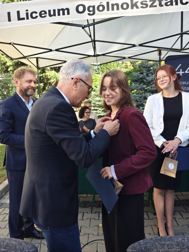 Starszy mężczyzna przypina niewielkie odznaczenie do bordowej marynarki młodej kobiety. Uczennica trzyma granatową teczkę, a obok stoją inni uczestnicy uroczystości.