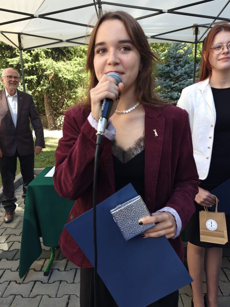 Młoda kobieta w bordowej marynarce mówi do mikrofonu. Trzyma granatową teczkę oraz niewielkie srebrne pudełko; za nią stoją inni uczestnicy wydarzenia.