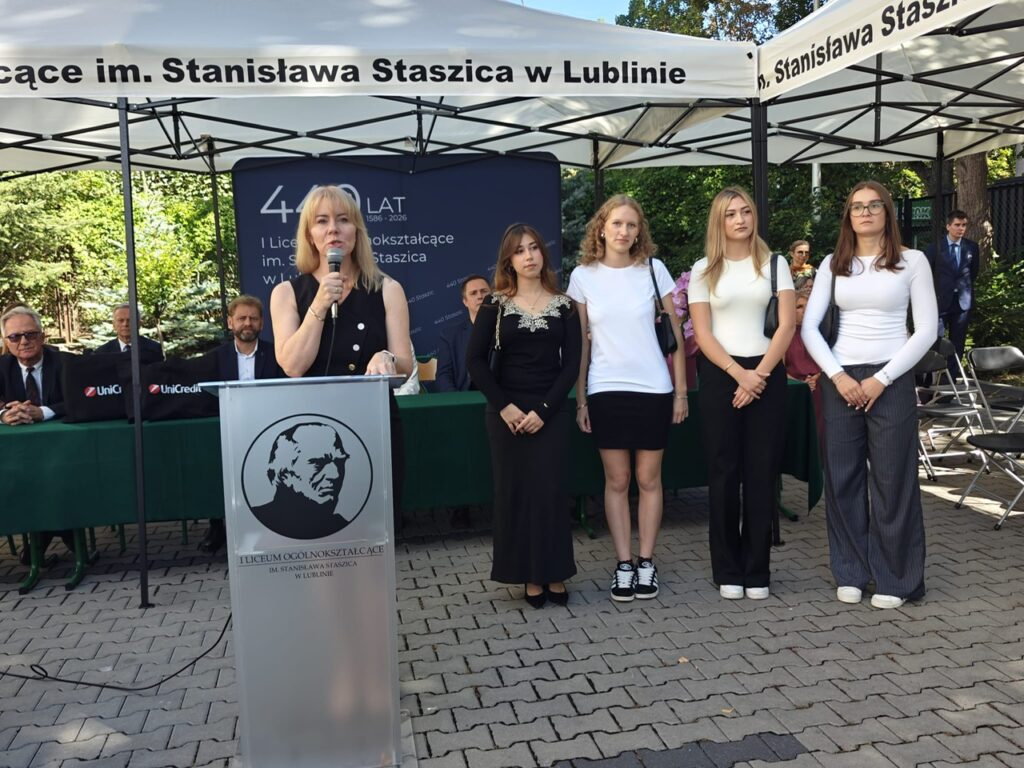 Kobieta w czarnej sukience przemawia do mikrofonu przy mównicy. Obok niej stoją cztery młode kobiety, a w tle widoczny jest jubileuszowy baner szkoły.