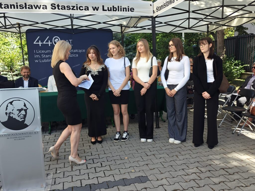 Kobieta w czarnej sukience stoi przed grupą pięciu młodych kobiet i przekazuje jednej z nich dokument. Obok znajduje się szkolna mównica.