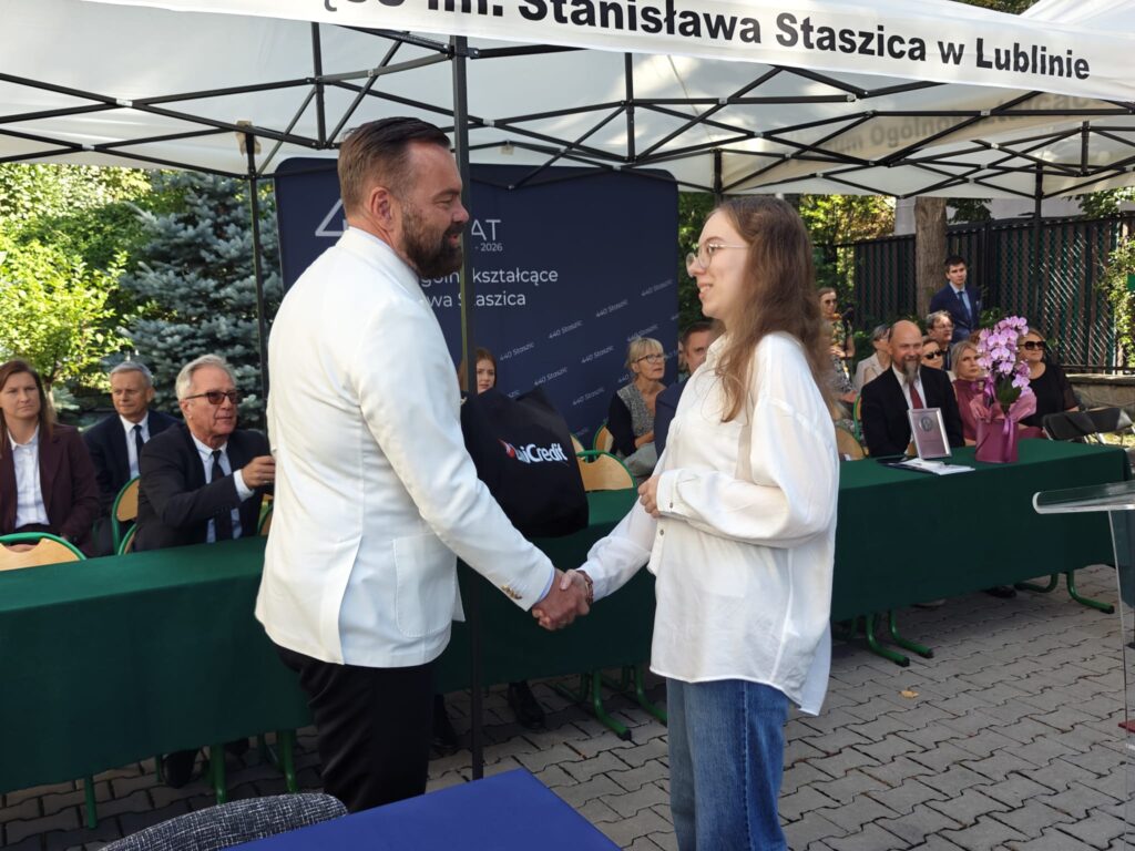 Mężczyzna w białej marynarce ściska dłoń uczennicy w białej koszuli i niebieskich spodniach. Za nimi znajduje się zielony stół oraz zaproszeni goście.