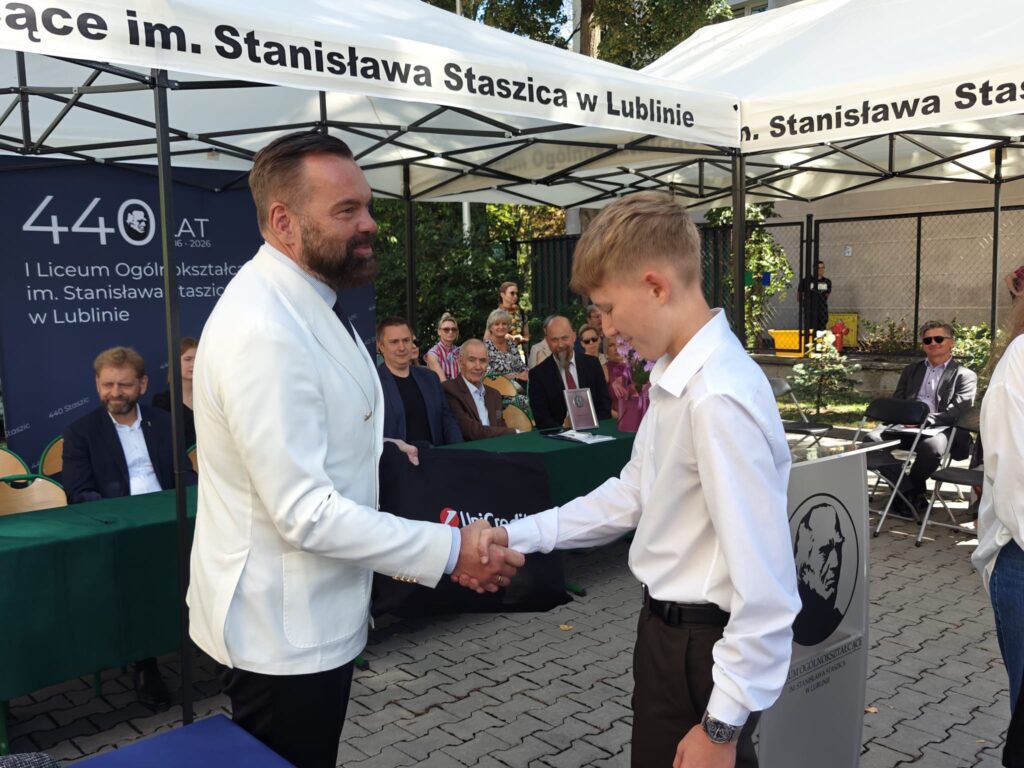 Mężczyzna w białej marynarce podaje rękę uczniowi w białej koszuli. W tle siedzą uczestnicy uroczystości.