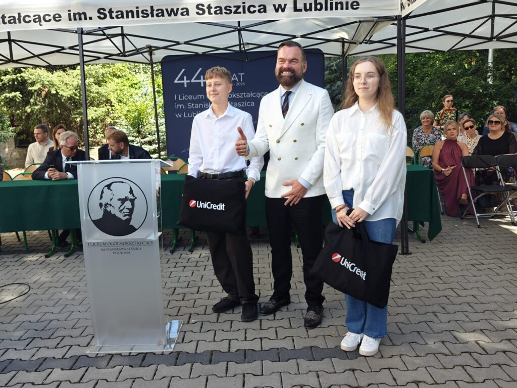 Mężczyzna w białej marynarce pozuje pomiędzy dwojgiem młodych ludzi. Uczniowie trzymają czarne torby z napisem „UniCredit”.