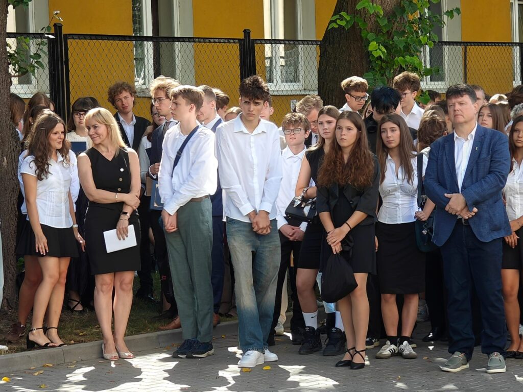 Duża grupa młodzieży i dorosłych stoi na szkolnym dziedzińcu. Uczniowie są ubrani głównie odświętnie, w białe koszule oraz ciemne spodnie i spódnice.