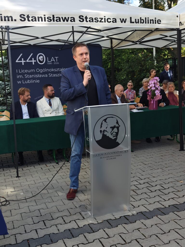 Mężczyzna w granatowej marynarce przemawia do mikrofonu przy przezroczystej mównicy z wizerunkiem patrona szkoły. W tle widoczny jest jubileuszowy baner „440 lat, 1586–2026, I Liceum Ogólnokształcące im. Stanisława Staszica w Lublinie”.