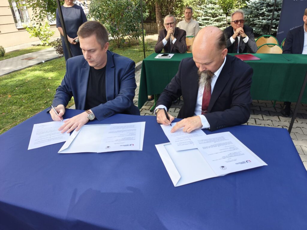 Dwóch mężczyzn siedzi przy stole nakrytym granatowym obrusem i podpisuje dokumenty. Za nimi, pod namiotem, przy zielonym stole siedzą uczestnicy uroczystości.