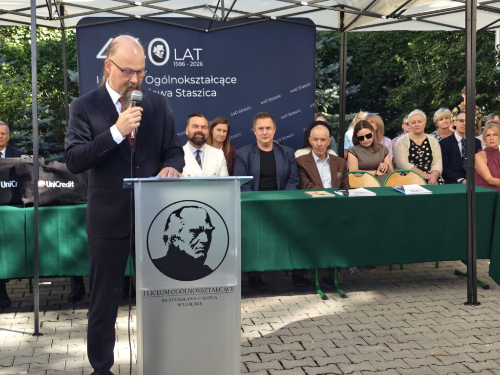 Mężczyzna w ciemnym garniturze przemawia przez mikrofon przy szkolnej mównicy. Za nim znajduje się plansza „440 LAT, 1586–2026” oraz liczna grupa uczestników siedzących przy zielonym stole.
