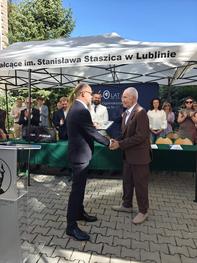 Dwaj mężczyźni podają sobie ręce podczas jubileuszowej uroczystości. Starszy mężczyzna w brązowym garniturze trzyma pamiątkową plakietę; w tle zgromadzeni uczestnicy biją brawo.