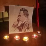 Na drewnianym krześle stoi czarno-biały portret mężczyzny z wąsami, przedstawionego z profilu, z głową opartą na dłoni. Przed fotografią ustawiono pięć zapalonych świeczek typu tealight. Ciepłe światło świec tworzy spokojną, nastrojową atmosferę.