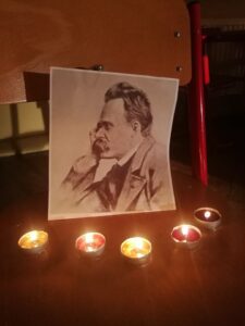 Na drewnianym krześle stoi czarno-biały portret mężczyzny z wąsami, przedstawionego z profilu, z głową opartą na dłoni. Przed fotografią ustawiono pięć zapalonych świeczek typu tealight. Ciepłe światło świec tworzy spokojną, nastrojową atmosferę.