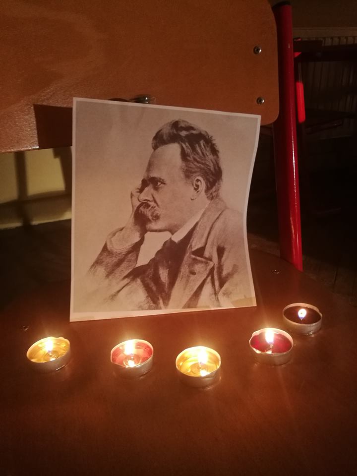 Na drewnianym krześle stoi czarno-biały portret mężczyzny z wąsami, przedstawionego z profilu, z głową opartą na dłoni. Przed fotografią ustawiono pięć zapalonych świeczek typu tealight. Ciepłe światło świec tworzy spokojną, nastrojową atmosferę.