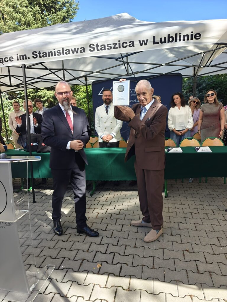 Starszy mężczyzna prezentuje uniesioną pamiątkową plakietę z medalem. Obok stoi mężczyzna w ciemnym garniturze, a zgromadzeni za zielonym stołem obserwują prezentację.