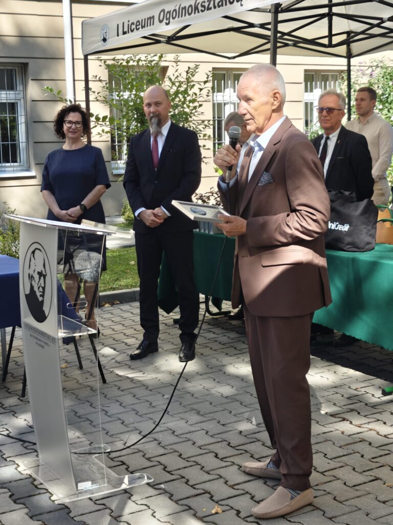 Starszy mężczyzna w brązowym garniturze przemawia przez mikrofon, trzymając pamiątkową plakietę. Obok stoją uczestnicy uroczystości, a po lewej znajduje się szkolna mównica.