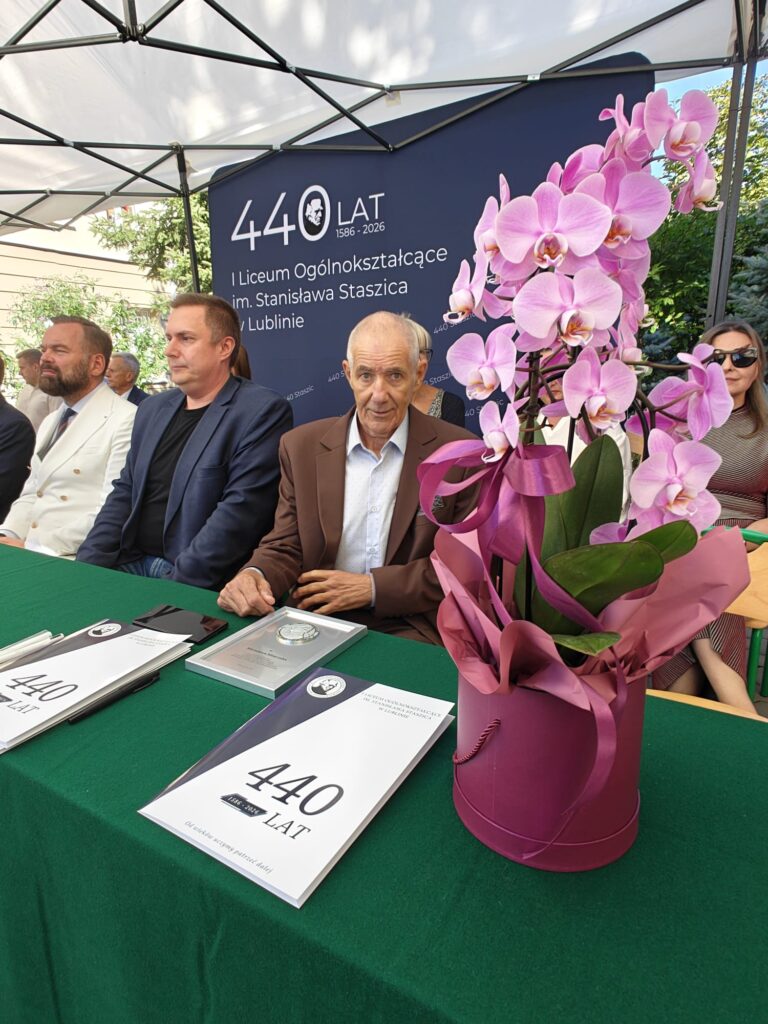 Uczestnicy jubileuszu siedzą przy zielonym stole. Na pierwszym planie znajduje się duża różowa orchidea w ozdobnym opakowaniu oraz publikacje z napisem „440 LAT, 1586–2026”.