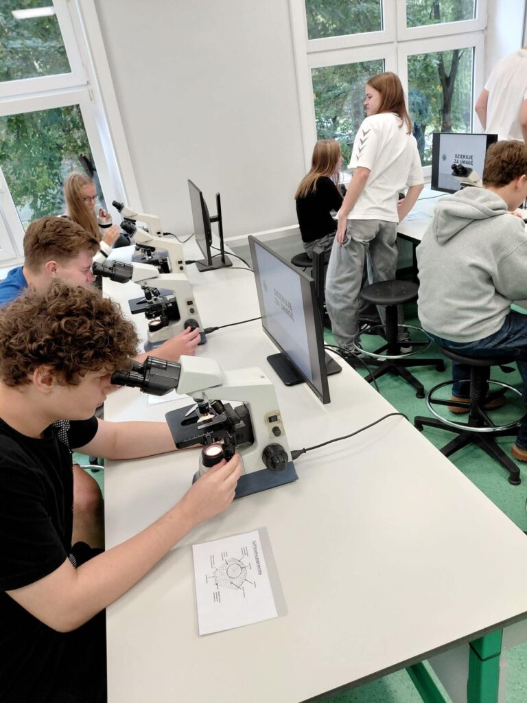 Uczniowie podczas praktycznych zajęć z mikroskopii. Na pierwszym planie uczeń obserwuje preparat przez mikroskop i reguluje urządzenie. Obok znajdują się kolejne stanowiska z mikroskopami, monitorami oraz materiałami dydaktycznymi.