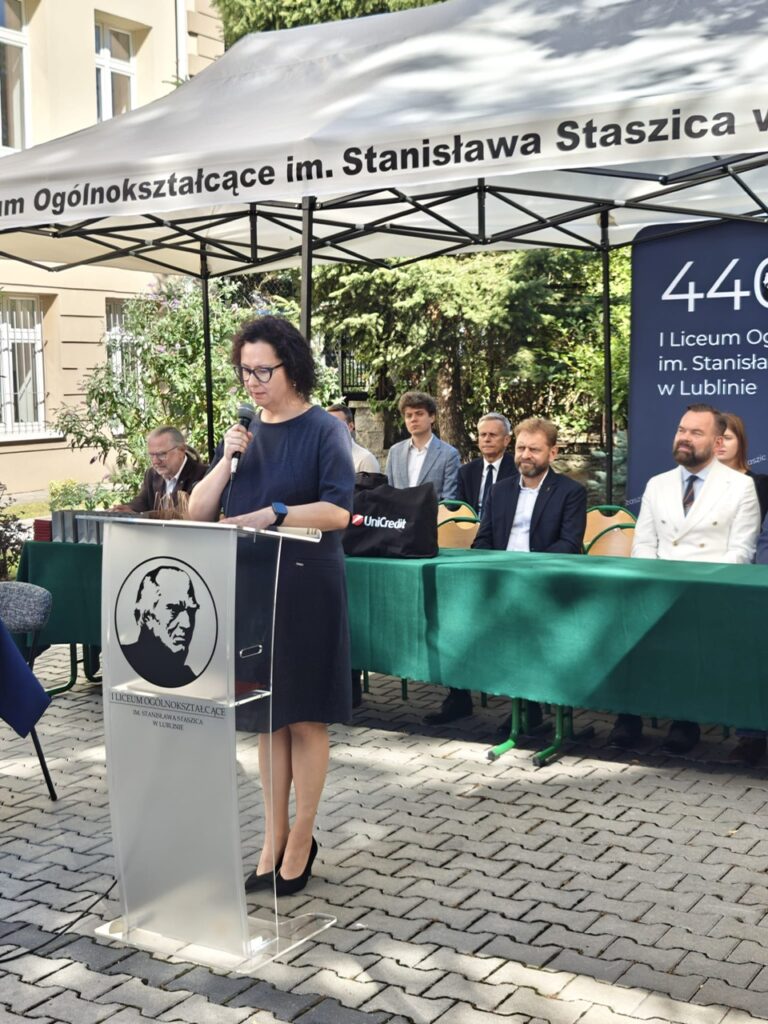 Kobieta w granatowej sukience przemawia do mikrofonu przy przezroczystej mównicy z symbolem szkoły. Za nią siedzą uczestnicy jubileuszu, a nad nimi widoczny jest napis z nazwą I Liceum Ogólnokształcącego im. Stanisława Staszica.