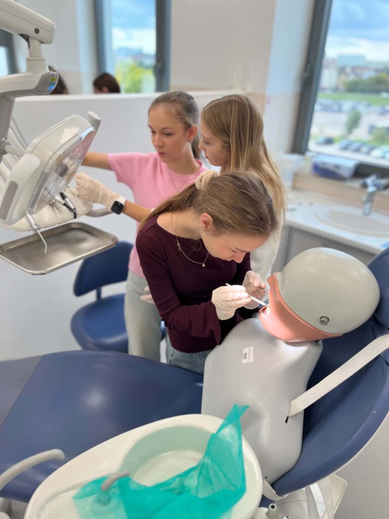 Trzy uczestniczki ćwiczą przy fotelu stomatologicznym. Jedna z nich wykonuje czynności przy modelu pacjenta, korzystając z narzędzi dentystycznych, a pozostałe obserwują.