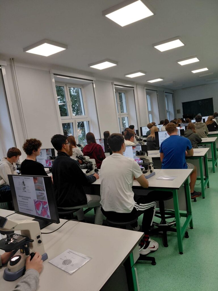 Pracownia biologiczna podczas zajęć. Uczniowie siedzą przy długich stołach wyposażonych w mikroskopy i monitory komputerowe. Na ekranach widoczne są materiały związane z obserwacjami mikroskopowymi. Sala jest jasna, z dużymi oknami i zieloną podłogą.