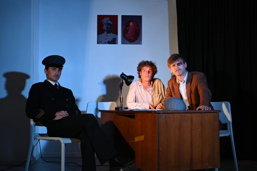 Trzej młodzi mężczyźni podczas sceny teatralnej. Po lewej siedzi mężczyzna w ciemnym mundurze i czapce, z nogą założoną na nogę. Za drewnianym biurkiem znajdują się dwaj pozostali: jeden w jasnej koszuli, drugi w brązowej marynarce. Obaj pochylają się do przodu i patrzą poza kadr. Na biurku stoją czarna lampa i szara czapka, a chłodne i ciepłe światło mocno modeluje twarze oraz rzuca cienie na ścianę.