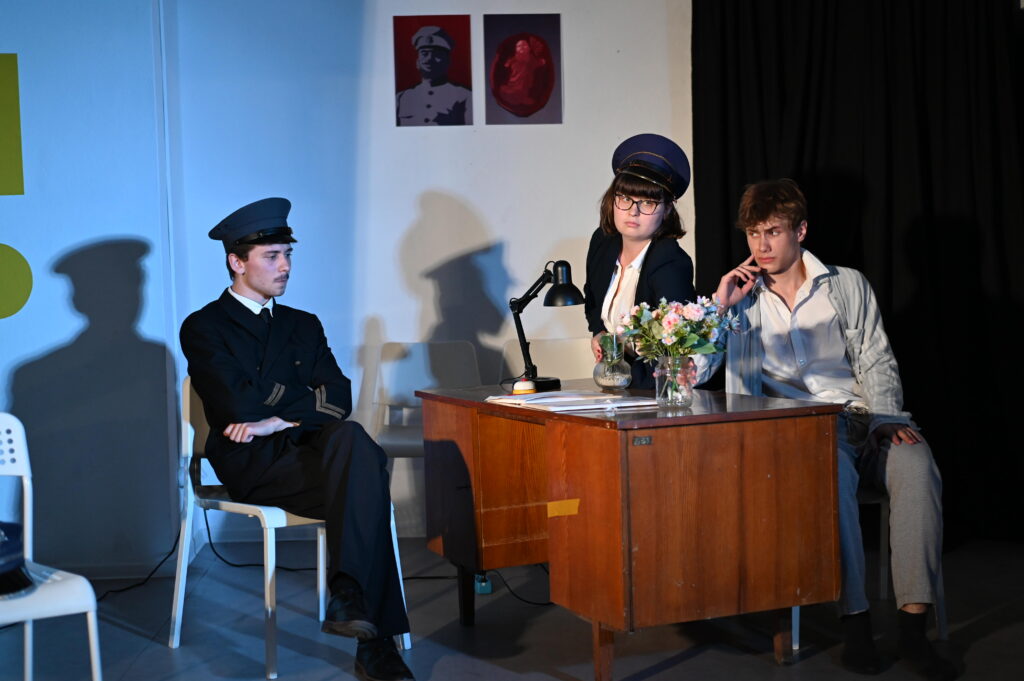 Trzy osoby podczas sceny teatralnej przy drewnianym biurku. Po lewej siedzi mężczyzna w ciemnym mundurze i czapce. Za biurkiem stoi kobieta w okularach, ciemnym żakiecie i mundurowej czapce, a obok siedzi młody mężczyzna w jasnej koszuli, opierający dłoń o twarz. Na biurku stoją lampa oraz dwa wazony z kolorowymi kwiatami.