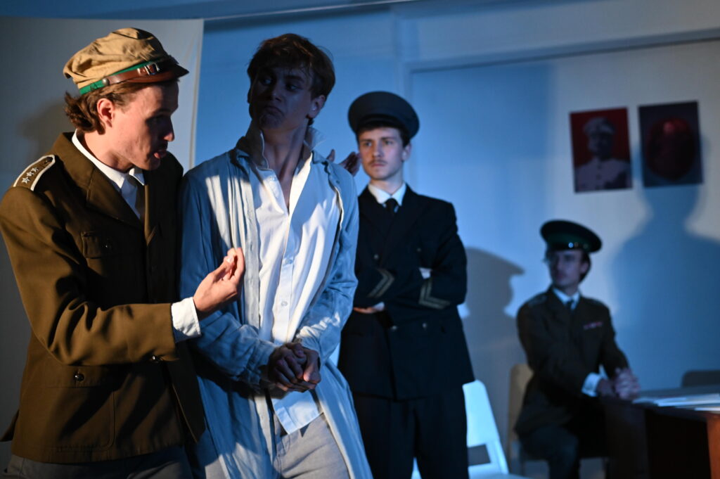 Zbliżenie sceny teatralnej. Mężczyzna w brązowo-zielonym mundurze i jasnej czapce gestykuluje podczas rozmowy z młodym mężczyzną w jasnym stroju, który trzyma dłonie razem i spogląda na rozmówcę. W głębi stoją lub siedzą dwaj kolejni mężczyźni w ciemnych mundurach. Chłodne, niebieskie oświetlenie nadaje scenie poważny charakter.