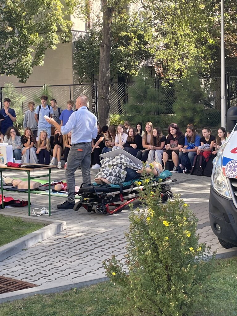 Prowadzący szkolenie stoi przy noszach z manekinem i zwraca się do siedzącej przed nim grupy młodzieży. Obok znajdują się manekiny do nauki resuscytacji oraz stół ze sprzętem szkoleniowym. Z prawej strony widoczny jest fragment ambulansu.