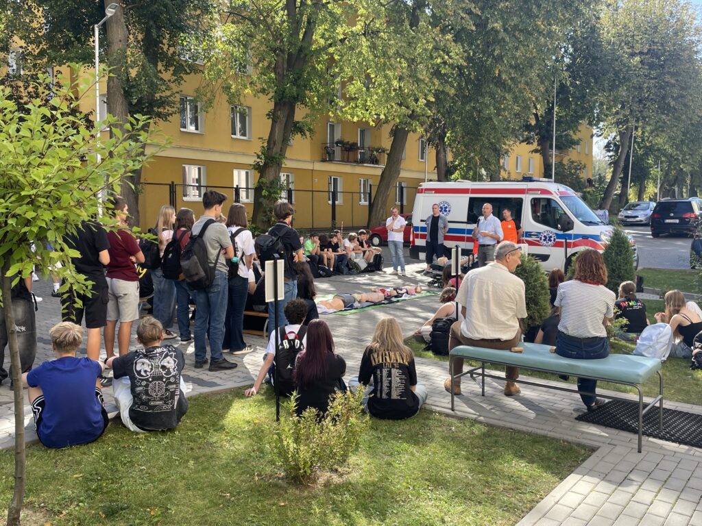 Szeroki widok szkolnego dziedzińca podczas zajęć z pierwszej pomocy. Liczna grupa uczniów siedzi i stoi wokół stanowiska demonstracyjnego. Przy ambulansie znajdują się prowadzący, a na matach pośrodku ułożono manekiny szkoleniowe.