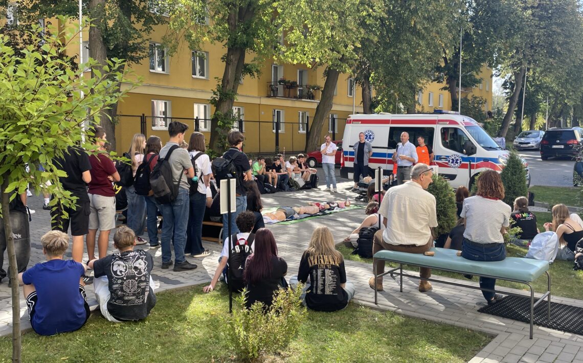 Szeroki widok szkolnego dziedzińca podczas zajęć z pierwszej pomocy. Liczna grupa uczniów siedzi i stoi wokół stanowiska demonstracyjnego. Przy ambulansie znajdują się prowadzący, a na matach pośrodku ułożono manekiny szkoleniowe.