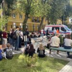 Szeroki widok szkolnego dziedzińca podczas zajęć z pierwszej pomocy. Liczna grupa uczniów siedzi i stoi wokół stanowiska demonstracyjnego. Przy ambulansie znajdują się prowadzący, a na matach pośrodku ułożono manekiny szkoleniowe.