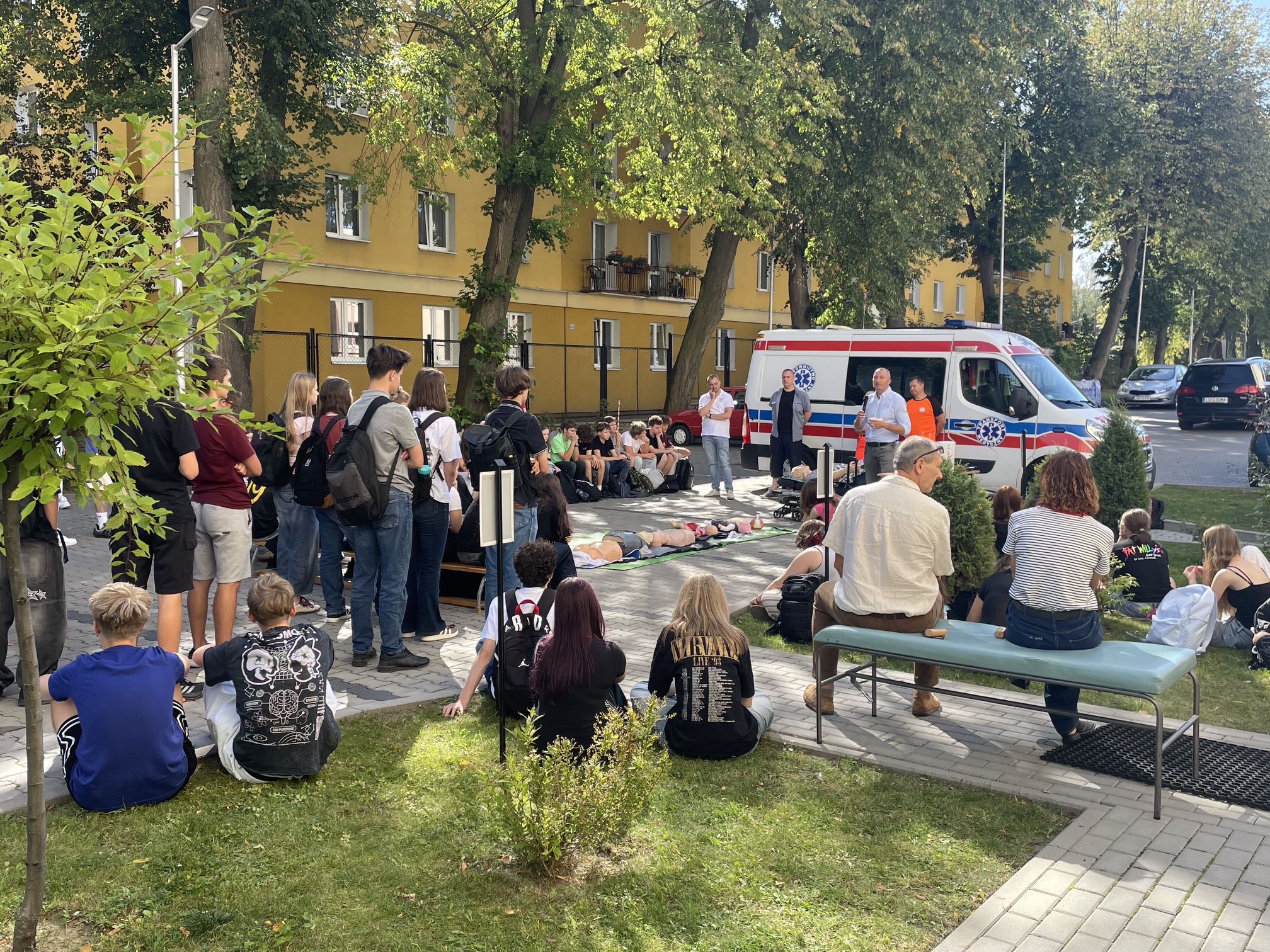 Szeroki widok szkolnego dziedzińca podczas zajęć z pierwszej pomocy. Liczna grupa uczniów siedzi i stoi wokół stanowiska demonstracyjnego. Przy ambulansie znajdują się prowadzący, a na matach pośrodku ułożono manekiny szkoleniowe.