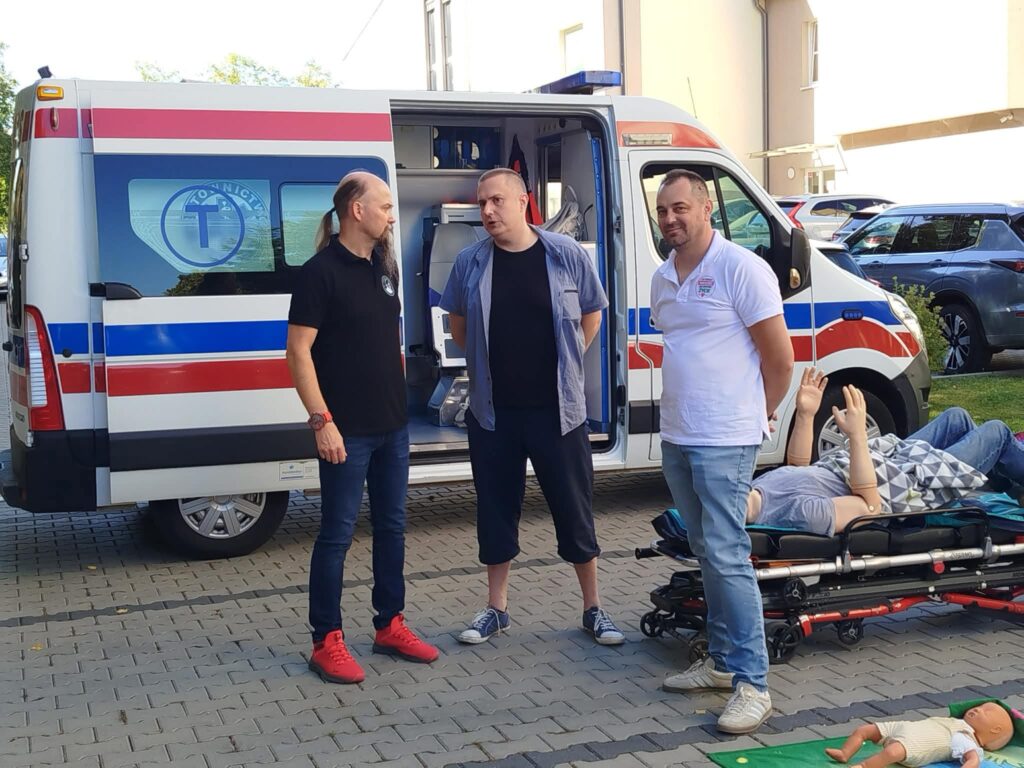 Trzech mężczyzn stoi przed otwartym ambulansem oznaczonym jako transport medyczny. Obok nich na noszach znajduje się manekin szkoleniowy z uniesionymi rękami. Na ziemi widoczny jest również mniejszy manekin do ćwiczeń pierwszej pomocy.