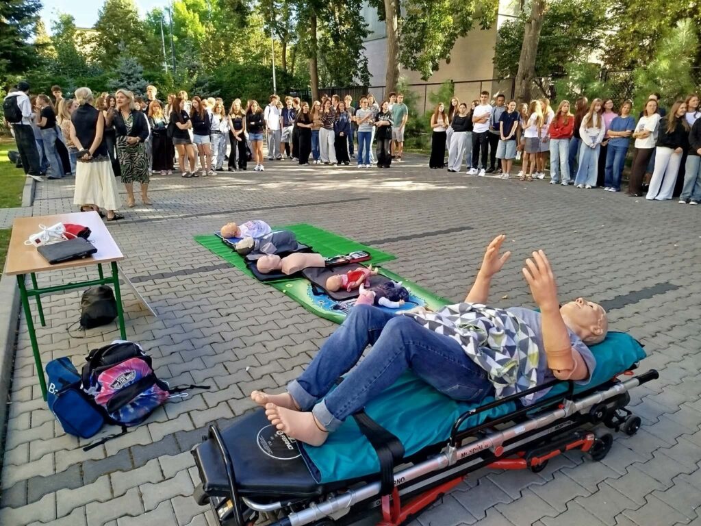 Uczniowie stoją w dużym półokręgu na dziedzińcu, obserwując stanowisko do nauki pierwszej pomocy. Na matach leżą manekiny do ćwiczeń resuscytacji, a najbliżej aparatu znajduje się pełnowymiarowy manekin umieszczony na noszach.