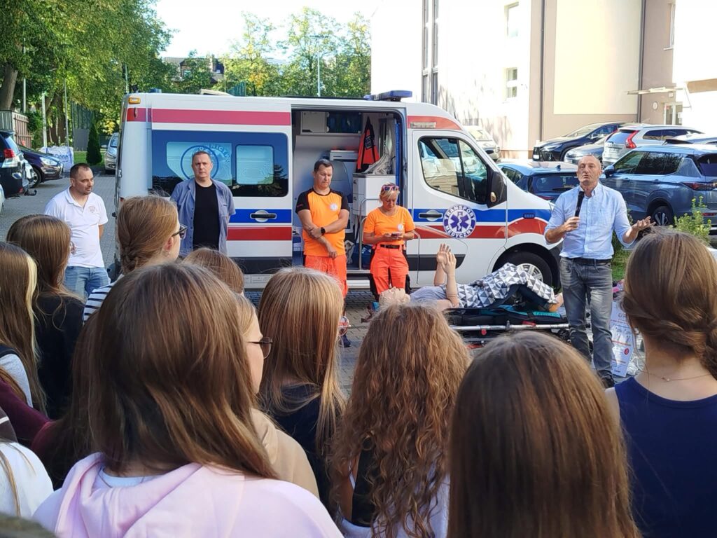 Pokaz ratownictwa medycznego na szkolnym dziedzińcu. Przed otwartym ambulansem stoją ratownicy oraz prowadzący szkolenie. Na noszach leży manekin szkoleniowy, a na pierwszym planie zgromadzona młodzież obserwuje demonstrację.