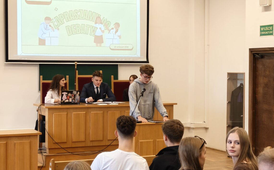 Zbliżenie na uczestnika debaty w szarej bluzie, stojącego przy mównicy z mikrofonem i korzystającego z przygotowanych materiałów. Za nim przy stole prezydialnym siedzą młodzi organizatorzy. Nad nimi wyświetlona jest prezentacja „Zapraszamy do debaty” z datą 25 września 2025.