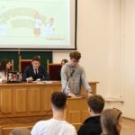 Zbliżenie na uczestnika debaty w szarej bluzie, stojącego przy mównicy z mikrofonem i korzystającego z przygotowanych materiałów. Za nim przy stole prezydialnym siedzą młodzi organizatorzy. Nad nimi wyświetlona jest prezentacja „Zapraszamy do debaty” z datą 25 września 2025.