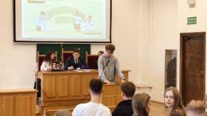 Zbliżenie na uczestnika debaty w szarej bluzie, stojącego przy mównicy z mikrofonem i korzystającego z przygotowanych materiałów. Za nim przy stole prezydialnym siedzą młodzi organizatorzy. Nad nimi wyświetlona jest prezentacja „Zapraszamy do debaty” z datą 25 września 2025.