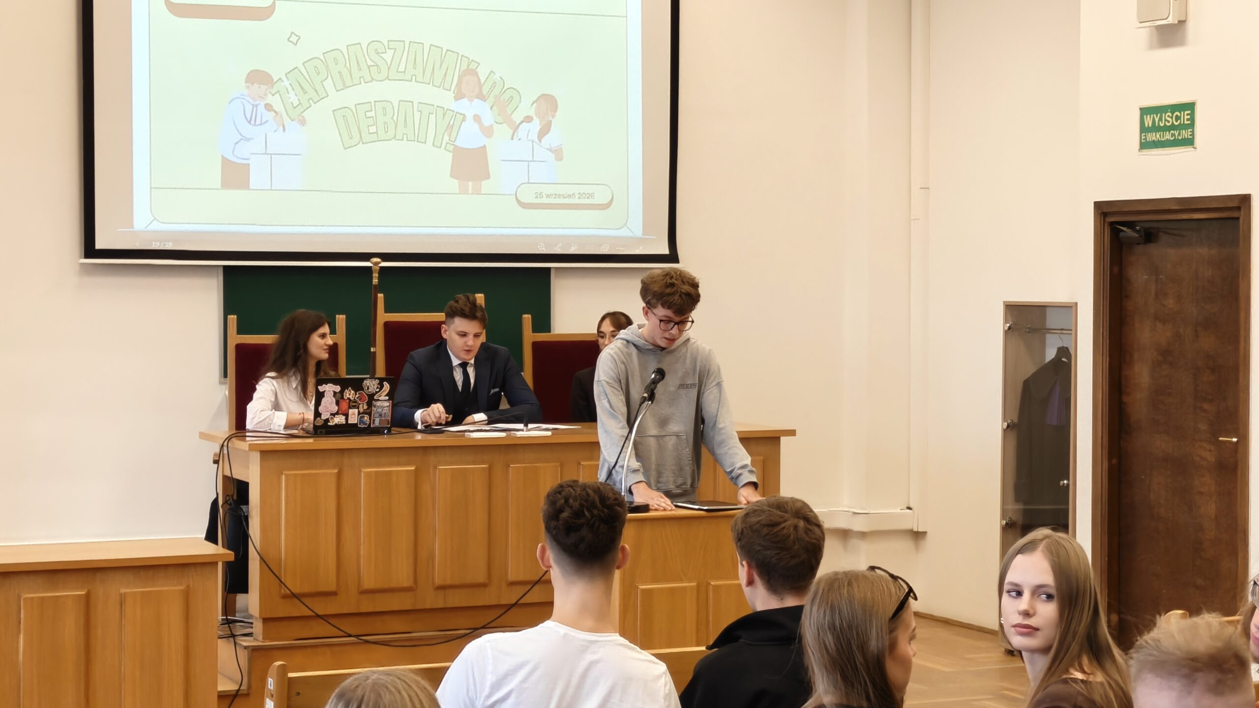 Zbliżenie na uczestnika debaty w szarej bluzie, stojącego przy mównicy z mikrofonem i korzystającego z przygotowanych materiałów. Za nim przy stole prezydialnym siedzą młodzi organizatorzy. Nad nimi wyświetlona jest prezentacja „Zapraszamy do debaty” z datą 25 września 2025.