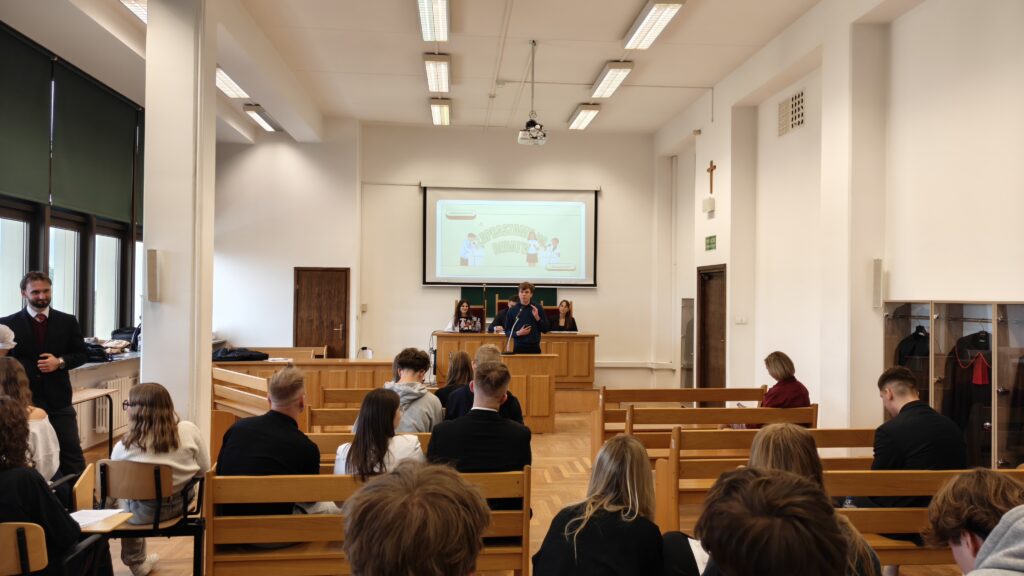 Ta sama aula podczas debaty. Przy mównicy stoi młody mężczyzna, który przemawia do siedzącej przed nim młodzieży. W tle widoczny jest ekran z prezentacją „Zapraszamy do debaty”.