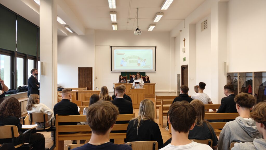 Sala przypominająca aulę lub salę rozpraw, wypełniona młodzieżą siedzącą na drewnianych ławach. Z przodu młoda kobieta przemawia przy mównicy, a za nią siedzą organizatorzy. Na ekranie wyświetlona jest prezentacja „Zapraszamy do debaty”.