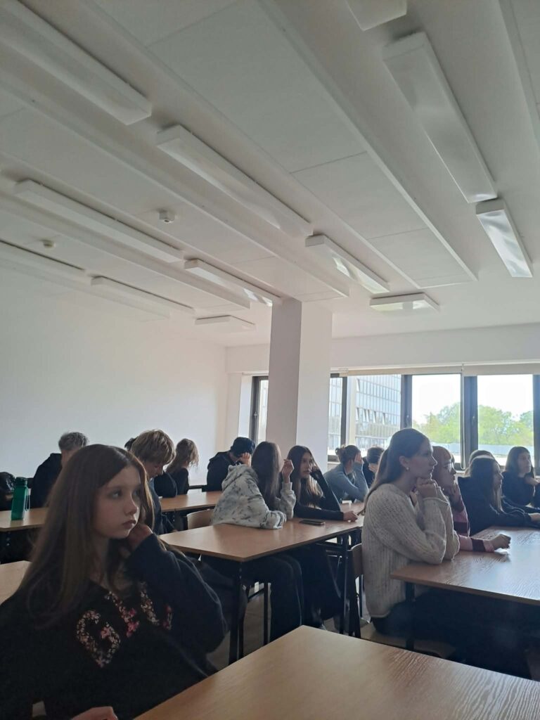 Grupa młodzieży siedzi przy stołach w jasnej sali wykładowej i uczestniczy w zajęciach. Uczniowie są zwróceni w stronę niewidocznego na zdjęciu prowadzącego.