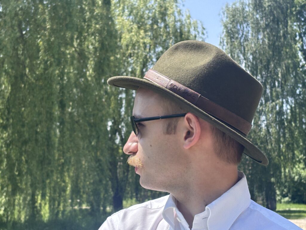 Mężczyzna w białej koszuli, ciemnych okularach i zielonym kapeluszu z brązowym paskiem, przedstawiony z profilu na tle zielonych, liściastych drzew w słoneczny dzień.