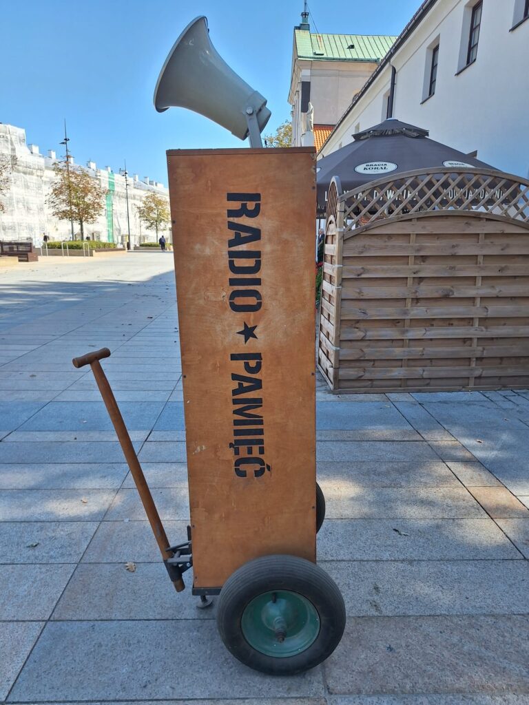 Mobilna instalacja „RADIO ★ PAMIĘĆ” – wysoka drewniana skrzynia na dwóch kołach, z dużym szarym megafonem umieszczonym na górze. Urządzenie stoi na miejskim placu.