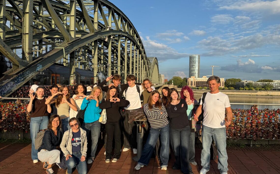 Grupowe zdjęcie młodzieży i opiekunów podczas wycieczki. Grupa pozuje na tle dużego stalowego mostu i rzeki, przy barierze gęsto pokrytej kolorowymi kłódkami. Część uczniów układa dłonie w kształt serca. Zdjęcie wykonano w ciepłym świetle późnego popołudnia.