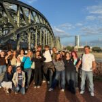 Grupowe zdjęcie młodzieży i opiekunów podczas wycieczki. Grupa pozuje na tle dużego stalowego mostu i rzeki, przy barierze gęsto pokrytej kolorowymi kłódkami. Część uczniów układa dłonie w kształt serca. Zdjęcie wykonano w ciepłym świetle późnego popołudnia.