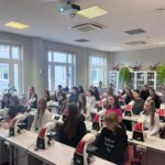 Uczniowie siedzą w kilku rzędach przy stanowiskach wyposażonych w mikroskopy. Wszyscy są zwróceni w stronę prowadzącego zajęcia, znajdującego się poza kadrem. W tle stoją przeszklone szafy laboratoryjne oraz duże rośliny.