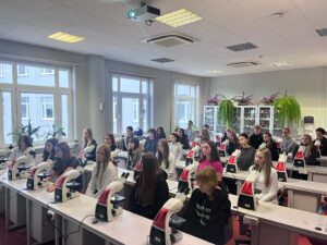 Uczniowie siedzą w kilku rzędach przy stanowiskach wyposażonych w mikroskopy. Wszyscy są zwróceni w stronę prowadzącego zajęcia, znajdującego się poza kadrem. W tle stoją przeszklone szafy laboratoryjne oraz duże rośliny.