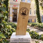 Drewniana statuetka z wizerunkiem Stanisława Staszica i napisem „I Liceum Ogólnokształcące im. Stanisława Staszica w Lublinie”, oznaczona numerem „#001”. Stoi na jasnej podstawie z datami „1586–2026”, w otoczeniu zielonego bluszczu, przed budynkiem szkoły.