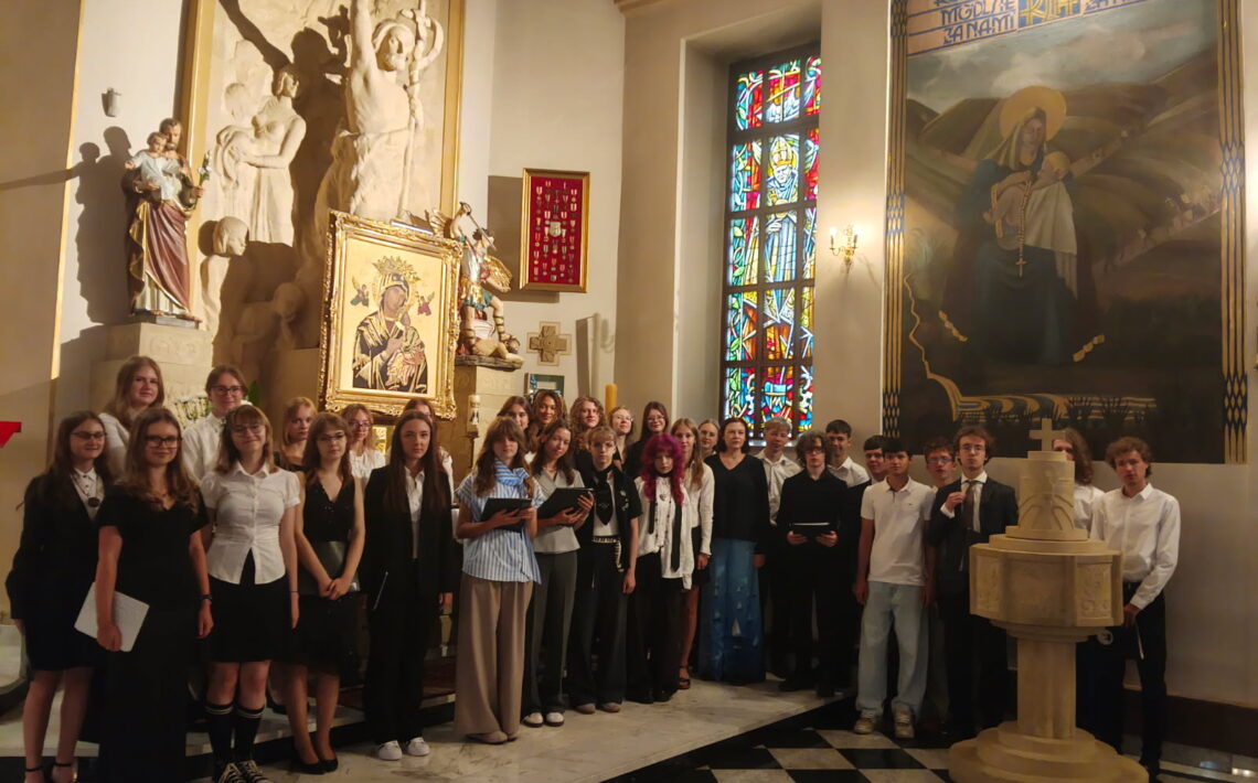 Grupa około 30 młodych osób pozuje do wspólnego zdjęcia we wnętrzu kościoła. Uczniowie stoją przed ołtarzem, część trzyma nuty lub śpiewniki. W tle widoczne są elementy wystroju sakralnego: kolorowy witraż, obrazy religijne, rzeźby oraz chrzcielnica. Wnętrze oświetla ciepłe światło słoneczne.