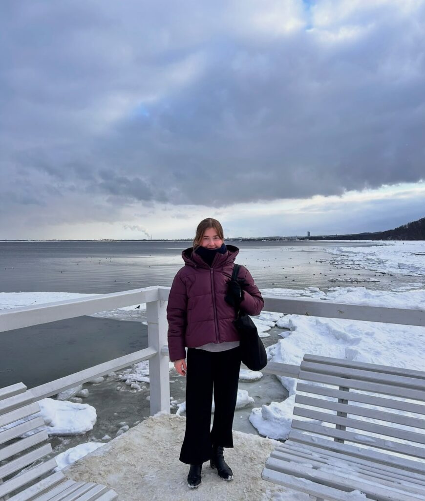 Młoda osoba stoi na zaśnieżonym nadmorskim pomoście, między jasnymi ławkami i białą barierką. Ma na sobie bordową kurtkę zimową, czarne spodnie, buty i szalik zasłaniający dolną część twarzy. W tle rozciąga się spokojne, częściowo skute lodem morze oraz ośnieżony brzeg. Nad krajobrazem dominują ciężkie, szaroniebieskie chmury.
