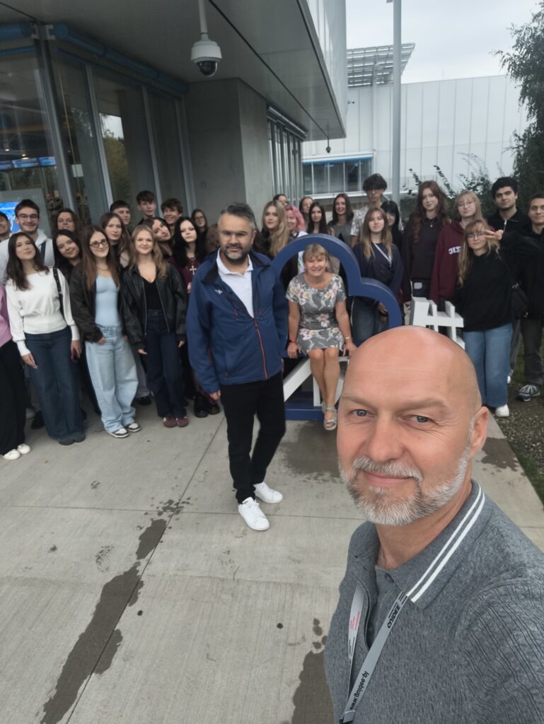 Grupowe selfie wykonane przed tym samym budynkiem. Na pierwszym planie, po prawej, znajduje się mężczyzna trzymający aparat lub telefon. Za nim pozuje liczna grupa młodzieży i dorosłych. Centralnie przy przestrzennym znaku siedzi kobieta w szarej sukience w kwiaty. Zdjęcie ma swobodny, pamiątkowy charakter.